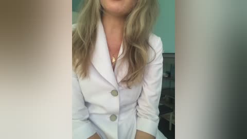 Media: A video of a woman with long, wavy blonde hair, wearing a white blazer with two silver buttons and a gold necklace. The background is blurred, featuring a light blue wall and indistinct objects.