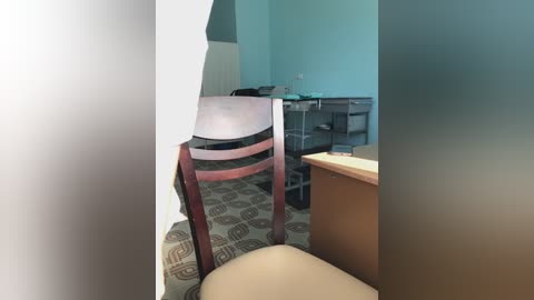 Media: Video of a sparsely furnished room with light blue walls, a wooden chair with a brown seat, beige carpet, and a desk with a computer monitor in the background.