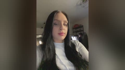 Media: A video of a young woman with long, straight black hair, fair skin, and a white top, looking down in a dimly lit room with shelves in the background.