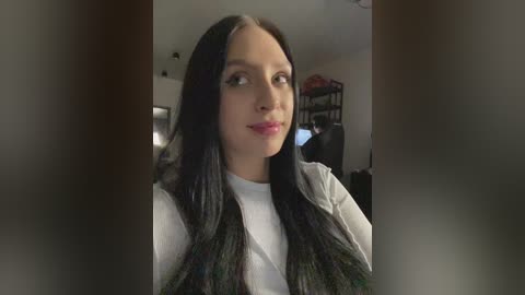 Video of a young woman with long, straight black hair and light skin, wearing a white shirt, indoors with blurred background.