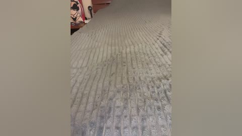 Video of a textured, light gray carpet with a ribbed pattern, partially visible wooden furniture in the background, and a blurred, dark-colored wall.