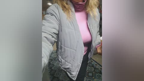 Media: Video of a woman with blonde hair, wearing a light gray quilted jacket over a pink turtleneck sweater and black pants. She stands in a room with a patterned rug and a blurred background.