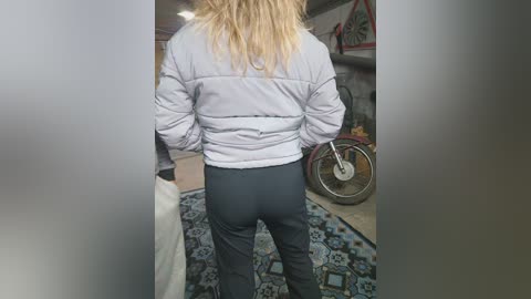 Media: Video of a person with long blonde hair, wearing a light grey puffy jacket and tight black pants, standing in a cluttered room with a patterned rug, a fan, and shelves in the background.