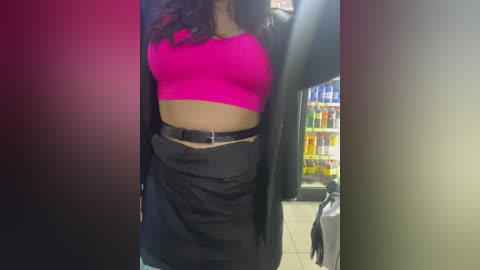 Media: Video of a woman with medium skin tone, wearing a bright pink crop top, black high-waisted shorts, and a black blazer, standing in a convenience store with colorful soda displays in the background.