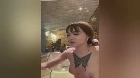 Media: Video of a nude, fair-skinned woman with short brown hair and a butterfly tattoo on her chest, sitting at a table in a dimly-lit room with exposed brick walls and soft lighting.