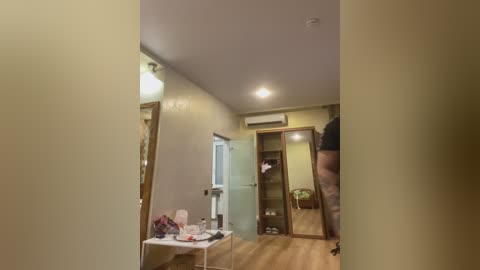 Media: Video of a small, narrow hallway with beige walls, wooden floor, open door, white table with cosmetics, mirror, and air conditioner on the ceiling.