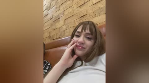 Video of a young Caucasian woman with short brown hair and pale skin, leaning on a brown leather bench in a public restroom. She has a slight smile, wearing a light-colored top, and the background features a textured yellow brick wall.