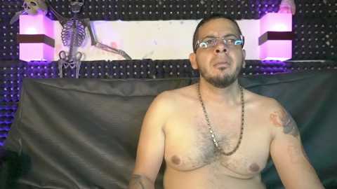 Video of a shirtless, light-skinned man with glasses and a goatee, sporting tattoos, standing in front of a wall with a skeletal art piece and black leather backdrop.