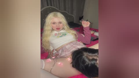 Media: Video of a young blonde woman with long hair, wearing a pink plaid skirt, white crop top, and white thigh-high stockings, lying on a pink bed, with a dark-haired person partially visible in the foreground.