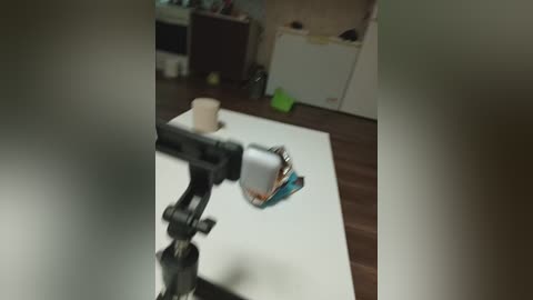 A blurry video of a table with a white sheet, a black tripod, and a small white object, possibly a camera, in the foreground. In the background, a messy room with dark wooden floors, shelves, and scattered objects.
