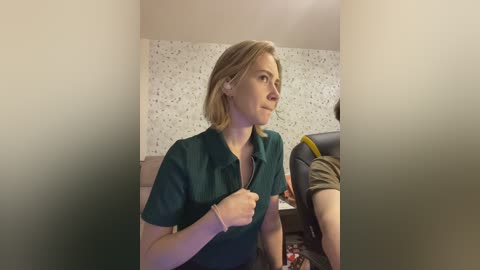 Video of a blonde woman in a teal shirt, seated on a beige chair in a room with a speckled wallpaper background and a green door.