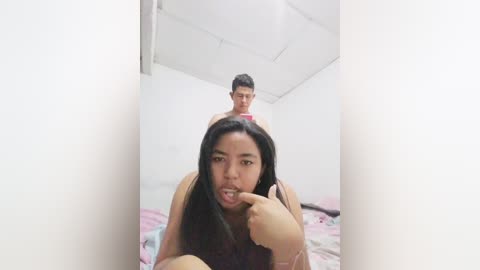 Video of an Asian woman with long black hair, wearing a beige top, playfully licking her finger while lying on a pink bed in a white room. A man stands behind her, partially visible.