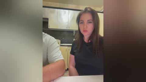 Media: Video of a Caucasian woman with long brown hair, wearing a black t-shirt, seated at a kitchen table, facing a man in a white t-shirt. Kitchen background with light wooden cabinets and a dark countertop.