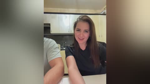 Media: Video of a smiling Caucasian woman with straight brown hair in a black t-shirt, sitting at a kitchen counter in a modern kitchen with light cabinets and dark countertops.