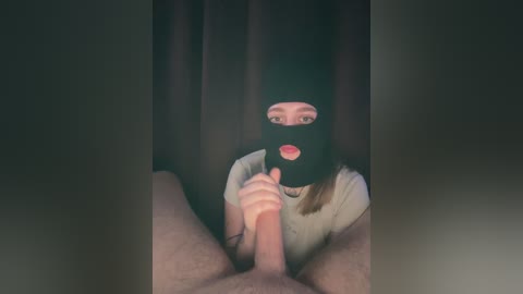 A video featuring a woman in a black balaclava and white top, gripping a circumcised penis, against a dark curtain background.