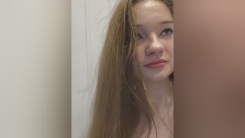 A video of a young Caucasian woman with long, wet, light brown hair, fair skin, and light makeup, partially obscured by a white wall and a person's arm. She looks towards the camera with a neutral expression.