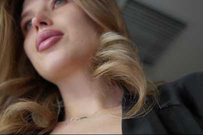 Media: Video of a close-up of a fair-skinned woman with long, wavy blonde hair, wearing a black top. Her expression is contemplative, and she has a delicate gold necklace.