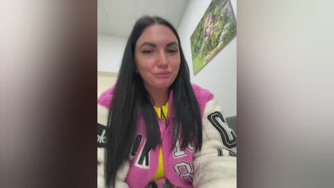 A video of a young woman with long black hair, fair skin, and a medium build, wearing a pink varsity jacket with white lettering, smiling. The background shows a white wall with a colorful painting of a forest scene.