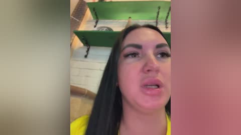 Video of a young woman with long black hair, wearing a bright yellow top, standing indoors next to green shelves with a chain hanging from the ceiling.