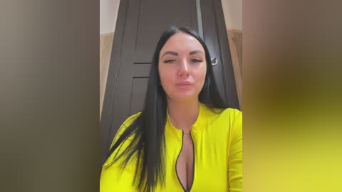 Video of a young woman with long black hair, wearing a bright yellow top, standing in front of a dark wooden door in a beige hallway.