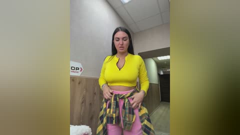 Video of a young woman with long black hair, wearing a bright yellow crop top and pink plaid pants, adjusting her shirt in a gym hallway with wooden walls and a \"Dorothy Perkins\" sign.