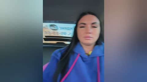 Video of a young woman with long black hair, wearing a blue hoodie, standing in a dimly lit room with a blurred poster in the background.