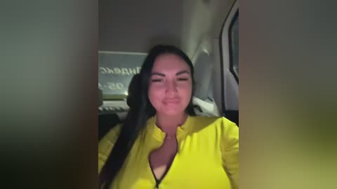 Video of a smiling, dark-haired woman in a yellow zip-up jacket, taken from a dimly lit car interior.