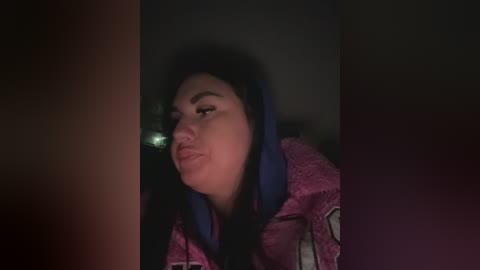 A dimly lit video of a young woman with long, dark hair, wearing a pink and blue hooded sweatshirt, puckering her lips in a playful kiss. The background is dark and out of focus, creating a moody, intimate atmosphere.