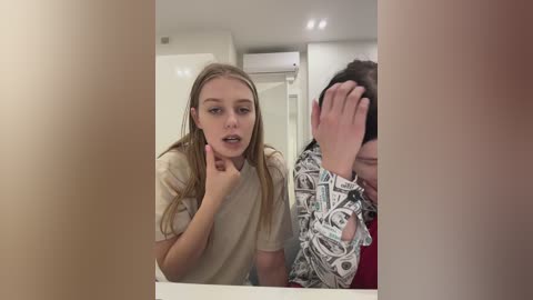 Media: Video of a young girl with long blonde hair, wearing a beige t-shirt, touching her chin. Behind her, a man with a patterned shirt is covering his face. They are in a modern bathroom with white cabinets and an air conditioner.