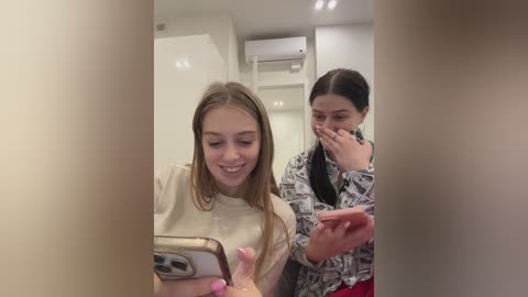 Media: Video of a young girl with long brown hair, smiling, holding a phone, in a modern, white-walled room with a ceiling air conditioner.