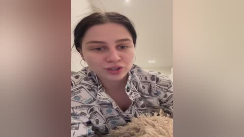 Media: A video of a young woman with fair skin, dark hair, and small hoop earrings, wearing a money-themed shirt, speaking into a microphone, in a beige room with soft lighting.