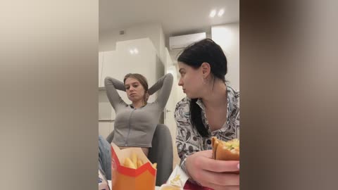 Media: Video of two women in a modern kitchen, one with a grey zip-up hoodie, hands behind head, the other in a patterned shirt, holding a burger.