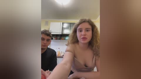 Media: A video featuring a young woman with curly brown hair in a beige bra, taking a selfie in a dimly-lit kitchen, with a young man in the background.