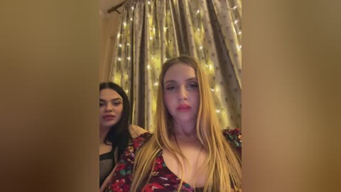 A video features two women; one with long blonde hair, wearing a red floral dress, and another with dark hair in a black top, standing behind her. They are in front of a curtain with fairy lights.