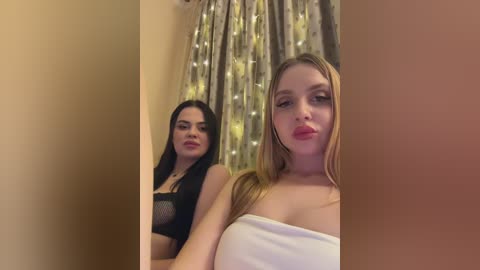 Video of two young women in a dimly-lit room. One with long black hair, wearing a black lace bra, stands behind another with long blonde hair, wearing a white strapless top.