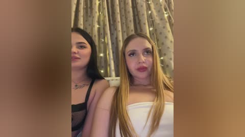 Video of two women, one with long straight hair and a white top, the other with long straight hair and a black mesh top, both with fair skin, posing against a curtain backdrop with soft lighting.