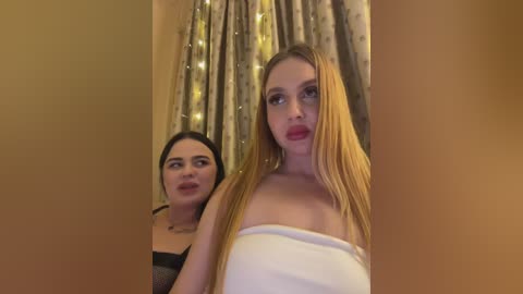 Video of two women in a dimly lit room; one with long blonde hair, white strapless dress, and red lipstick, standing; the other with dark hair, black top, and tan skin, in the background.