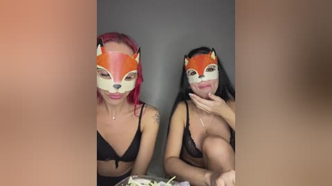 Video of two women with fair skin, wearing orange fox masks and black lingerie, sitting side-by-side against a gray wall, blurred background, intimate setting.