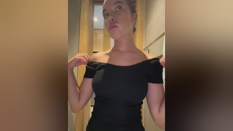 Media: Video of a fair-skinned woman with brown hair, wearing a black off-shoulder top, standing in a hallway with beige walls. She holds the straps of her top, looking serious.