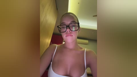 Media: Video of a fair-skinned woman with blonde hair, wearing black-framed glasses and a white tank top, standing in a dimly lit room with yellow walls and a visible air conditioner.