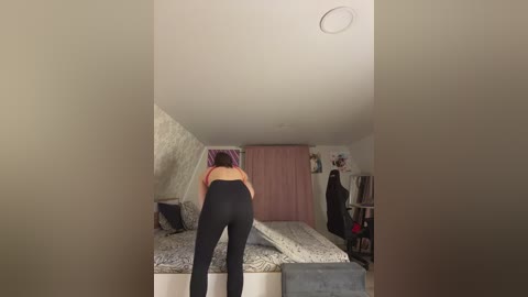 Media: Video of a woman in a small, cluttered bedroom with sloped ceiling, leaning over a bed, wearing a black bra and high-waisted pants, walls adorned with posters and clothes hanging on racks.