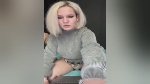 Media: Video of a blonde woman with a short bob haircut, wearing a grey sweater and patterned shorts, kneeling on a turquoise surface, against a dark background, capturing a candid, close-up selfie.