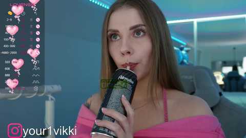 Media: Video of a young Caucasian woman with long, straight brown hair, light skin, wearing a pink off-shoulder top, drinking from a can, in a dimly lit room with blue LED lighting.