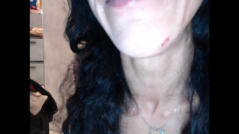 Video of a close-up of a person with long, curly black hair, fair skin, and a small facial scar, wearing a necklace. Background shows a kitchen with a white countertop and a black bag.