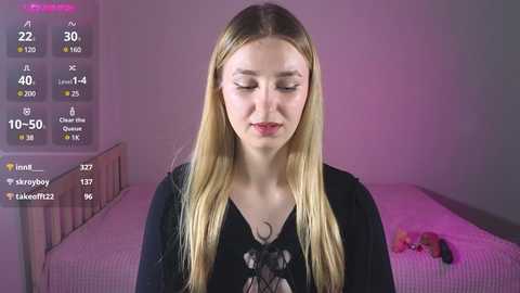 A video of a young, fair-skinned woman with long, blonde hair, wearing a black shirt, standing in a dimly lit bedroom with a bed and a pink wall.