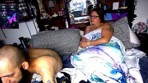 Video of a muscular, shirtless man lying face down on a couch with a tattooed woman sitting next to him, covered in a tie-dye blanket, in a cluttered, dimly lit living room.