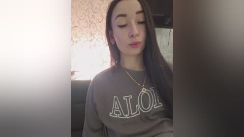 A video of a young woman with fair skin, long dark hair, and closed eyes, wearing a gray sweatshirt with \"ALOHA\" in white letters, seated in a dimly lit room with a white floral-patterned wallpaper.
