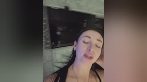 A video of a young woman with fair skin and long dark hair, lying on a bed, eyes closed, wearing a black bra.
