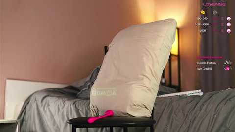 Video of a bed with gray bedding, a beige pillow, and a pink vibrator on a black stool, set against a pink wall with a warm, ambient light.