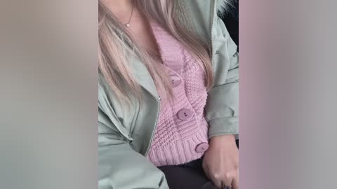 A video of a person with long, straight, platinum blonde hair, wearing a light green jacket over a pink knitted sweater with large buttons and a black skirt. The background is blurry.